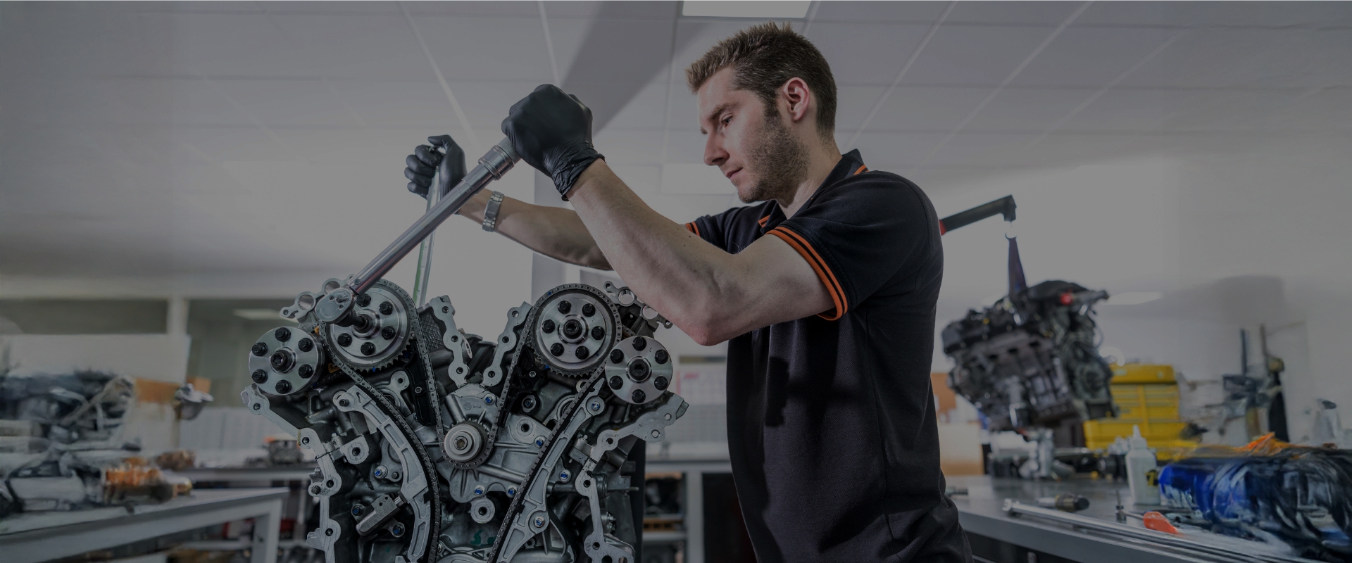 Ein Automechaniker in einer Werkstatt prüft Daten auf einem Tablet, während ein anderer Mechaniker auf einer Hebebühne an einem angehobenen Auto arbeitet.